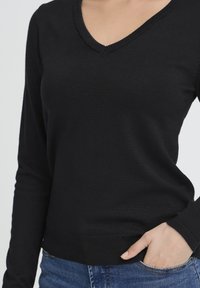 Mustast V-kaelusega kampsun pikkade varrukatega, kootud tekstuuriga ja soonilise äärega. Sobitatud siniste teksadega, rõhutades vabaaja sobivust.