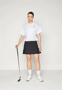 Hvit polo t-skjorte med korte ermer og krage, kombinert med et svart plisset skjørt til golf. Modellen holder en golfkølle og har på seg hvite sokker og joggesko.