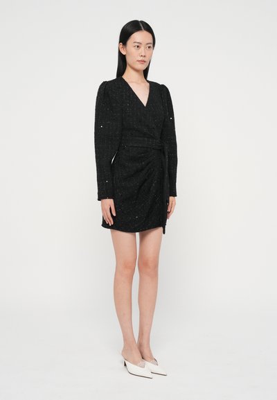 Melna apmetņa kleita no teksturēta auduma ar garām piedurknēm. Raksturīga sānu savērpšana un josta viduklim. Modele valkā baltas mules kurpes.