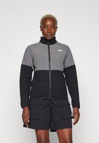Fleece jaka ar pelēku augšējo paneli un melno apakšējo daļu. Augsta apkakle, pilna rāvējslēdzēja aizdare un sānu kabatas. Valkāta ar melniem cargo šortiem.