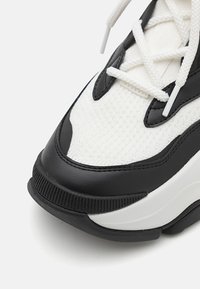 Svart og hvit sportsko med teksturert mesh-overdel, svarte lærdetaljer og kontrasterende gummisåle. Inkluderer hvite snørebånd.