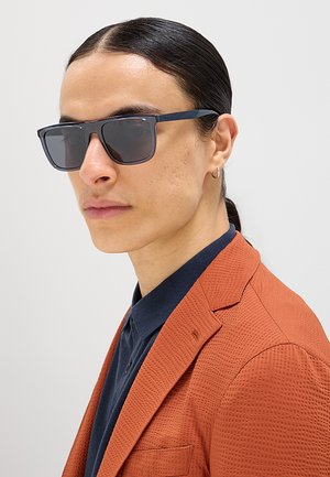Giovane uomo con lunghi capelli scuri legati dietro che indossa occhiali da sole neri, una giacca arancione strutturata e una camicia blu scuro.