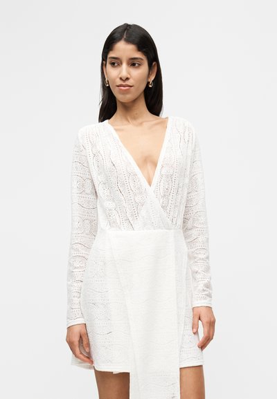 Λευκό φόρεμα από δαντέλα με μακριά μανίκια και βαθύ ντεκολτέ V. Διαθέτει περίπλοκα σχέδια και σχεδιασμό τύπου wrap με υφασμάτινο κάτω μέρος με υφή.