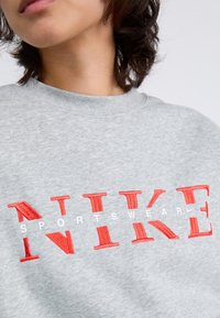 Sivi dukserica od pamučne mješavine, sa crvenim izvezenim "NIKE" logotipom i bijelim tekstom "SPORTSWEAR" ispod. Klasičan dizajn sa okruglim izrezom.