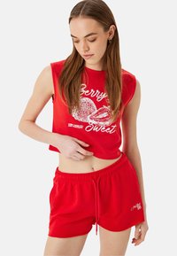 Top corto sin mangas rojo con texto gráfico blanco y diseño de fresa; combinado con pantalones cortos rojos a juego con cinturilla elástica y detalle de logo.