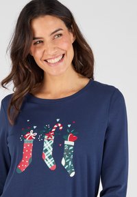 Top de manga larga azul marino con un gráfico festivo de tres medias decoradas en rojo, verde y blanco con corazones y bastones de caramelo.
