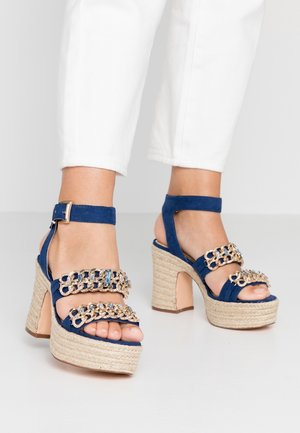 Pieds portant des sandales à talons hauts compensés bleues avec des détails en chaîne dorée et des brides à la cheville, associées à un pantalon blanc.