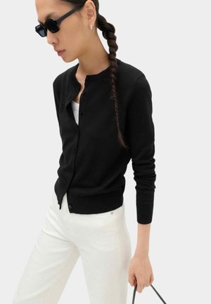 Frau trägt schwarzen, geknöpften Cardigan, weiße Hose und Sonnenbrille, hält schwarze Handtasche mit langem geflochtenem Haar.