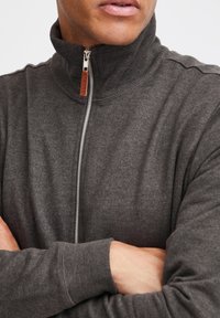 Donkergrijze zip-up sweatshirt met hoge kraag, ribgebreide manchetten en textiel. Voorzien van een zilveren rits en een bruin leren trekstik.