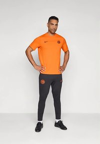 Vīrietis stāv ar rokām uz gurniem, ģērbies oranžā īsajās piedurknēs un melnā biksēs ar saskaņotiem oranžiem logo, pret vienkāršu gaišu fonu.