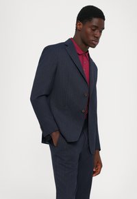Giovane uomo in un completo blu a righe con le mani in tasca, indossando una polo bordeaux su uno sfondo bianco semplice.