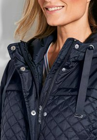 Zwarte gewatteerde jas van glanzende stof, met zilveren drukknoopsluitingen, ritsen en verstelbare banden. Heeft een hoge kraag en een textuurdesign.