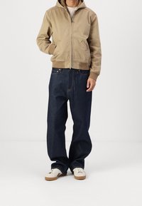 Beige zip-up hoodie with a hood, two side pockets; paired with dark blue denim wide-leg jeans and white sneakers with brown soles.