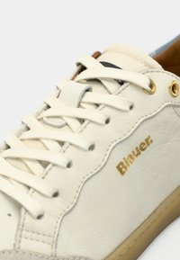 Λευκό δερμάτινο αθλητικό παπούτσι με χρυσές λεπτομέρειες, υφή επιφάνειας και επίπεδα κορδόνια. Διαθέτει διακριτικό σχέδιο και λαστιχένια σόλα. Λογότυπο στο πλάι.