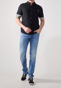 Camicetta nera a maniche corte con bottoni, con polsini blu a contrasto e una tasca marrone, abbinata a jeans azzurri chiari e sneakers nere.