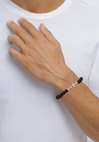 Svart lavastensarmband med ett silverkors som accent, bärs på en hand mot en vit t-shirtbakgrund.