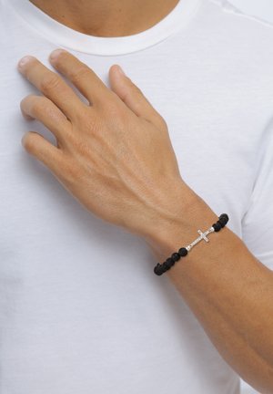 Black lava stone bracelet featuring a silver cross accent, worn on a hand against a white t-shirt background.