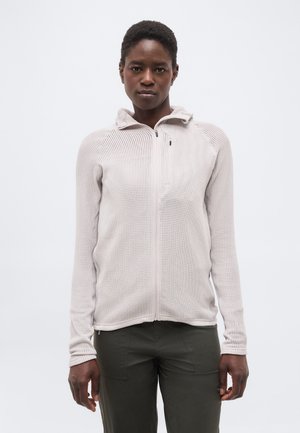 Persona que lleva una chaqueta beige clara con textura y cremallera, con cuello alto, y pantalones verde oliva oscuro, de pie contra un fondo claro y liso.