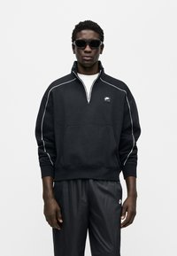 Μαύρο φούτερ με φερμουάρ στο μισό μέρος, λευκή πιπίλα και μικρό λογότυπο στο στήθος. Συνδυασμένο με μαύρο παντελόνι φόρμας, φορεμένο με γυαλιά ηλίου.