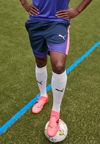 Puma Sportshorts in Marineblau mit pinken Akzenten und Muster, kombiniert mit weißen Socken mit schwarzem Logo und knalligen pinken Fußballschuhen auf einem Ball.