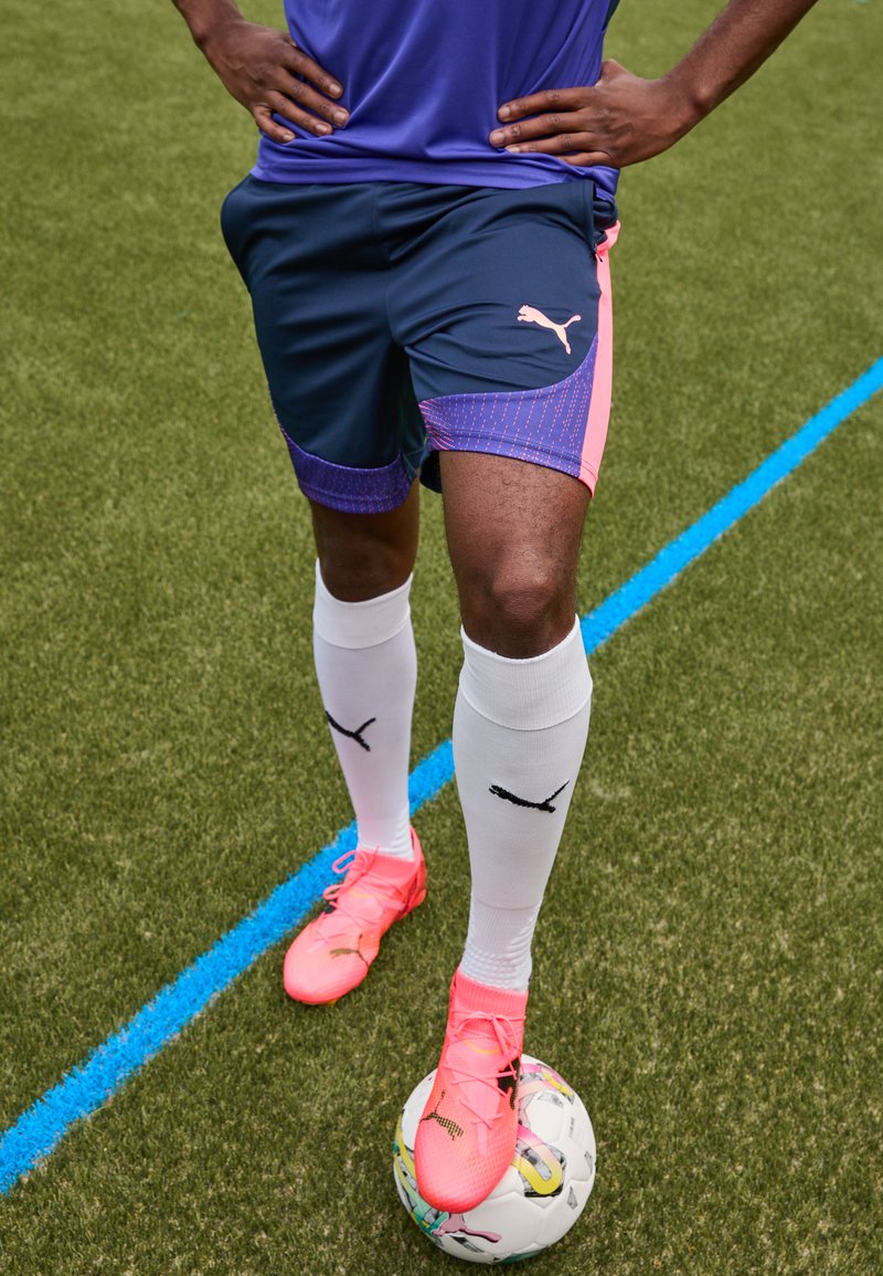Puma Sportshorts in Marineblau mit pinken Akzenten und Muster, kombiniert mit weißen Socken mit schwarzem Logo und knalligen pinken Fußballschuhen auf einem Ball.