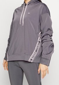 Gray sports hoodie with pink drawstrings and side accent details featuring a repeating logo pattern. Soft, smooth texture, relaxed fit.