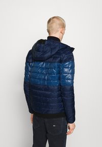 Donkerblauwe gewatteerde jas met capuchon, voorzien van horizontale banen in verschillende blauwtinten, gladde textuur en zwarte accenten.