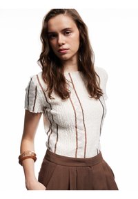 Biała bluzka o fakturowanej powierzchni z brązowymi pionowymi paskami, krótkimi rękawami i lekko falbaniastym wykończeniem; zestawiona z brązowymi spodniami o wysokim stanie.