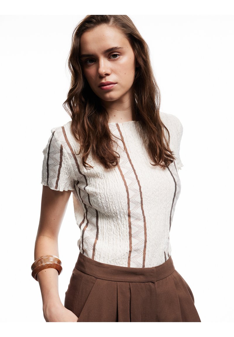 Biała bluzka o fakturowanej powierzchni z brązowymi pionowymi paskami, krótkimi rękawami i lekko falbaniastym wykończeniem; zestawiona z brązowymi spodniami o wysokim stanie.