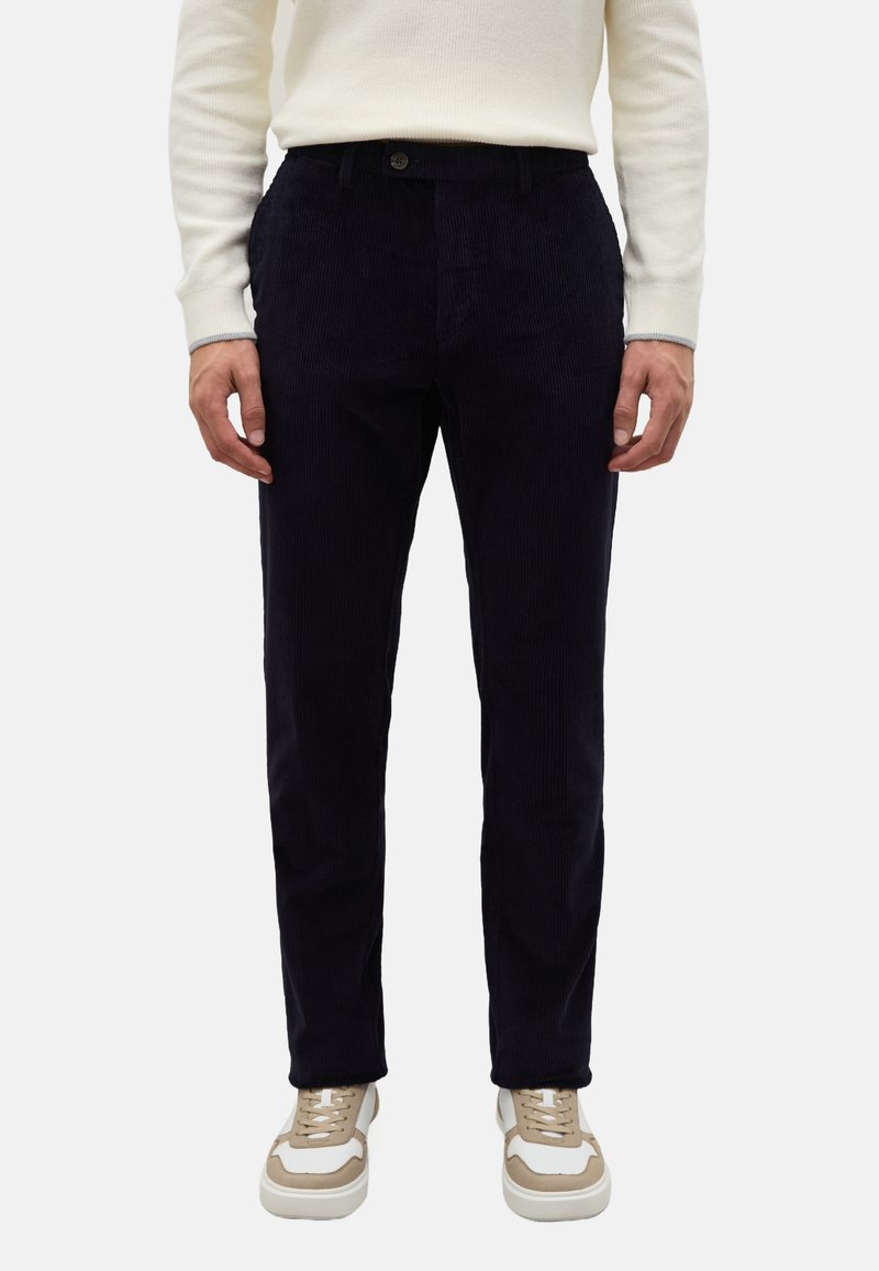 Marineblauwe corduroy broek met verticale geribbelde textuur, voorzien van een knoopsluiting en zijzakken, gecombineerd met beige sneakers.