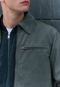 Veste grise avec un col structuré, poche zippée sur le devant et tissu texturé, superposée à un vêtement sombre zippé.