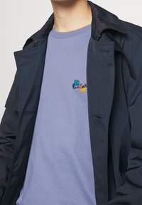Personne portant un t-shirt violet clair avec un petit texte de logo coloré et un manteau ouvert bleu marine foncé par-dessus.
