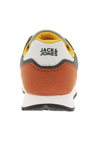 Vista trasera de una zapatilla con parte superior verde, talón de gamuza naranja, detalles blancos, suela negra y forro interior amarillo con logo.