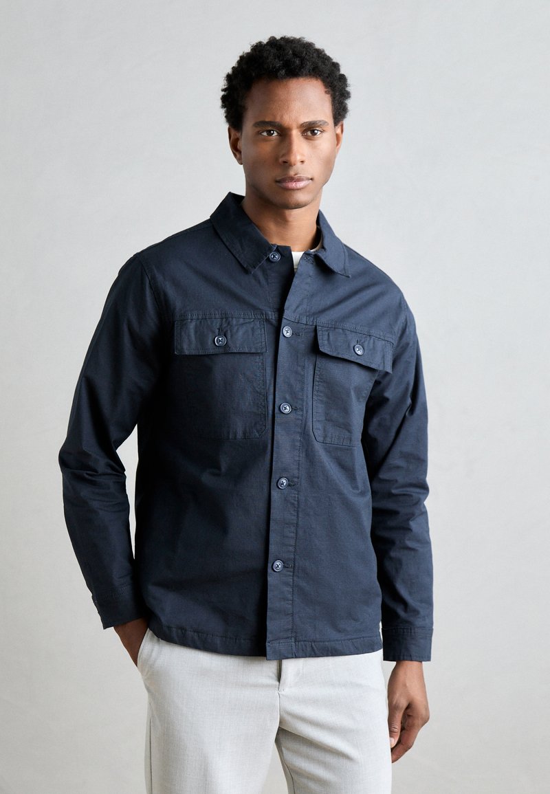 Camicia blu navy con colletto, due taschini sul petto con bottone, maniche lunghe e texture liscia. Adatta per un look casual.