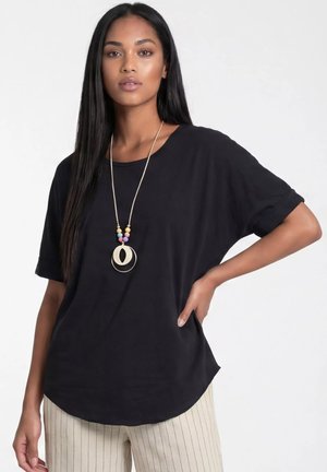 Kobieta o długich czarnych włosach nosi luźną czarną koszulkę, beżowe spodnie w cienkie prążki oraz długi naszyjnik z kolorowymi koralikami i geometrycznymi zawieszkami.