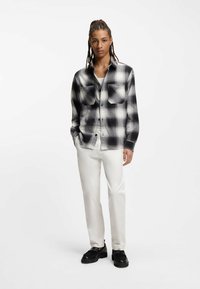 Μπλε πουκάμισο σε ασπρόμαυρο καρό με δύο τσέπες στο στήθος, σχεδιασμένο με κουμπιά, φορεμένο πάνω από λευκό τοπ και συνδυασμένο με λευκό παντελόνι και μαύρα παπούτσια.