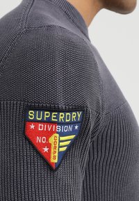 Grauer, gestrickter Pullover mit strukturierter Oberfläche; verfügt über ein bunt besticktes Patch auf der Schulter mit der Aufschrift "SUPERDRY DIVISION" und Sternen.