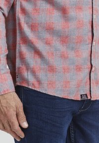 Rød og grå rutet flanellskjorte med lange ermer, knappedeling foran og et merke ved kanten. Kombinert med mørkeblå jeans.