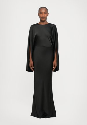 Mujer luciendo un vestido de satén negro de longitud completa con mangas anchas que presentan largas aberturas, de pie contra un fondo claro y sencillo.