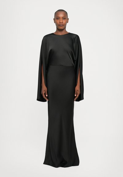 Woman wearing a floor-length black satin gown with wide sleeves featuring long slits, standing against a plain light background.