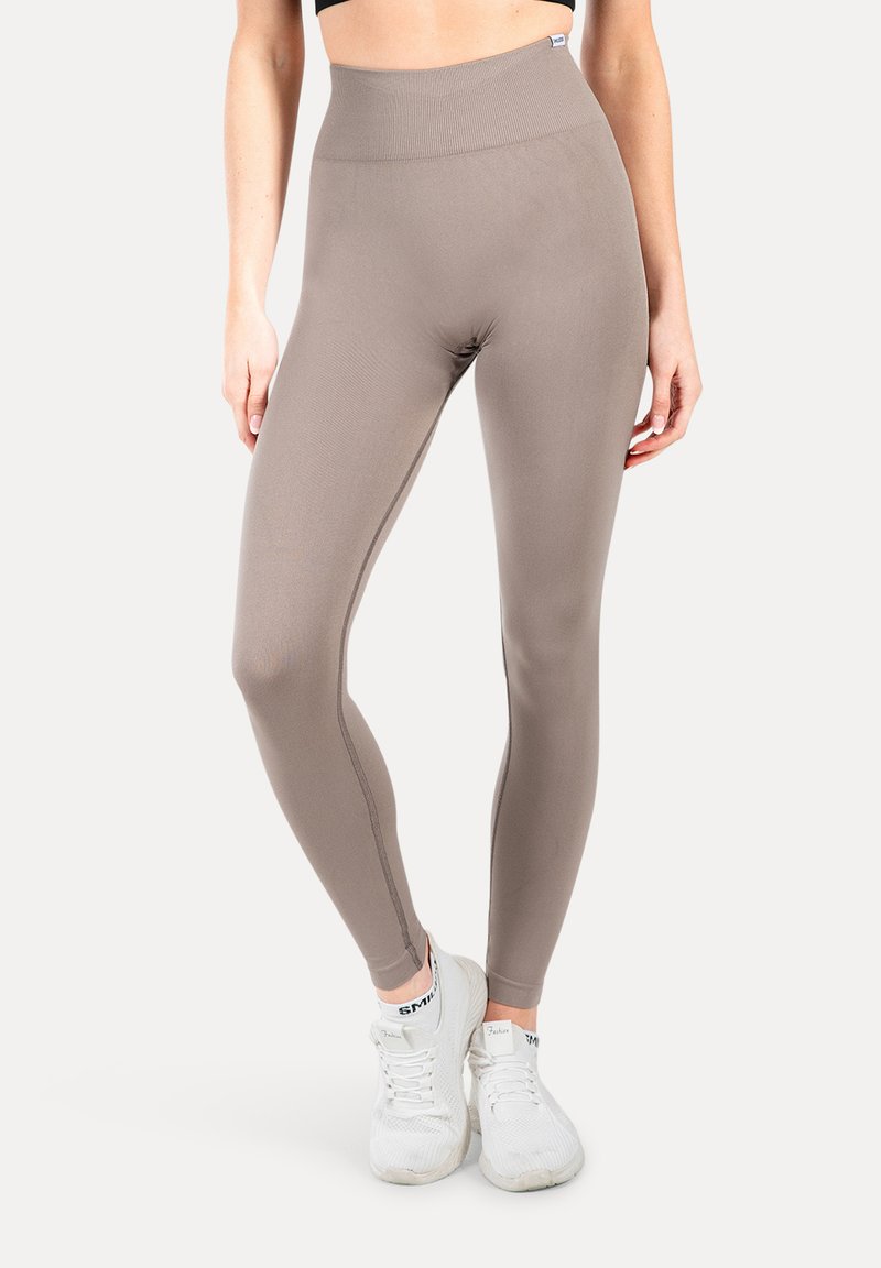 Leggings marroni a vita alta realizzati in materiale elastico con una texture liscia. Presentano cuciture a contrasto e sono indossati con scarpe da ginnastica bianche.