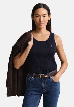 Femme aux longs cheveux foncés portant un débardeur bleu marine en maille torsadée, un jean bleu et une ceinture marron, tenant une veste brun foncé sur son épaule.