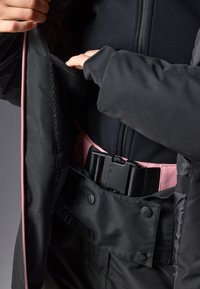 Svart jakke med glidelåslomme og en rosa belteakent. Innvendig har den et nettingfôr, og stoffet ser glatt og slitesterkt ut.