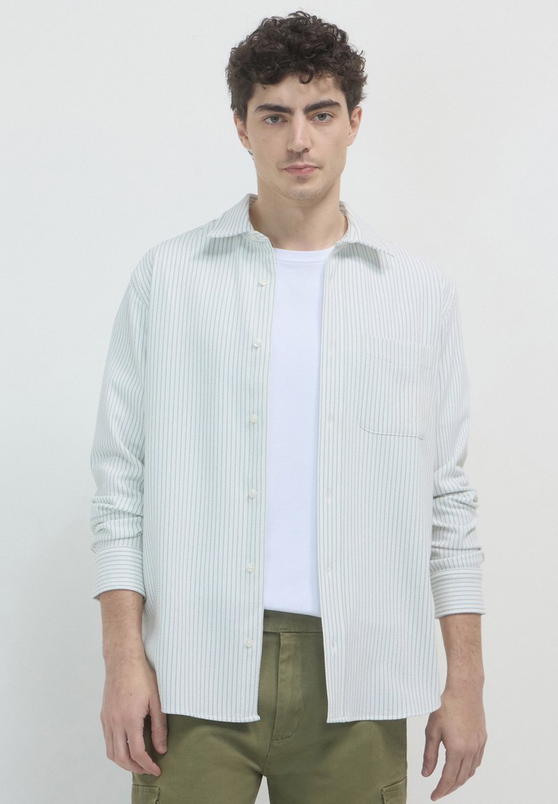 Un jeune homme aux cheveux bouclés porte une chemise blanche à rayures, sur un t-shirt blanc et un pantalon vert olive, devant un fond clair.
