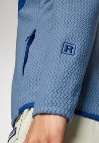 Lys blå fleecejakke med et teksturert sikksakkmønster, med blå trim og en liten brodert logo på ermet.