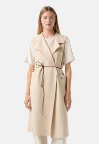 Beige Wollmischung-Weste mit tailliertem Kragen, einem Gürtel in der Taille, zwei vorderen Taschen und lockerer Passform, kombiniert mit einem hellen Oberteil.