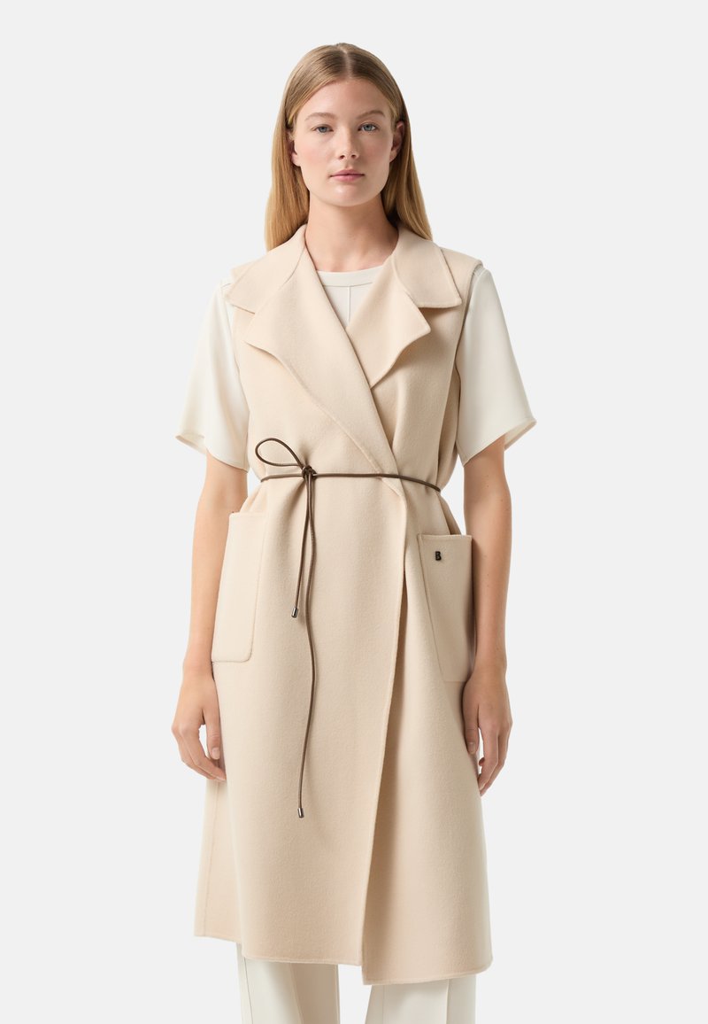 Beige Wollmischung-Weste mit tailliertem Kragen, einem Gürtel in der Taille, zwei vorderen Taschen und lockerer Passform, kombiniert mit einem hellen Oberteil.