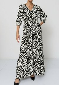 Robe maxi en imprimé zèbre noir et blanc. Présente un design croisé, des manches bouffantes, une ceinture à nouer et un ourlet à volants en couches.