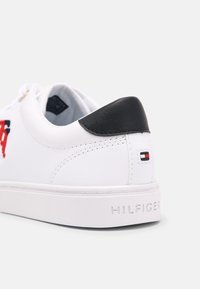 White leather sneaker with a black heel accent, embossed logo, and multicolored branding on the side. Flat white rubber sole.