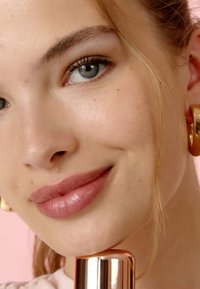 Zbliżenie twarzy o jasnej skórze, błyszczących różowych ustach i czystych oczach. Złote kolczyki w kształcie kół oraz metaliczny różowo-złoty wkład trzymany w ręku.